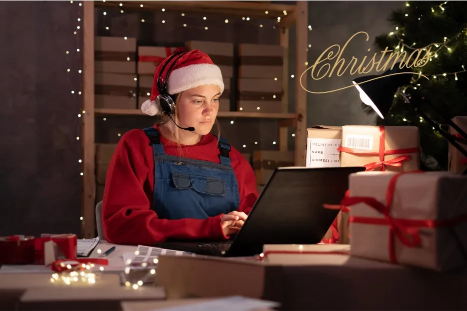 Woman in Santa hat with headset uses laptop surrounded by Christmas gifts and lights.