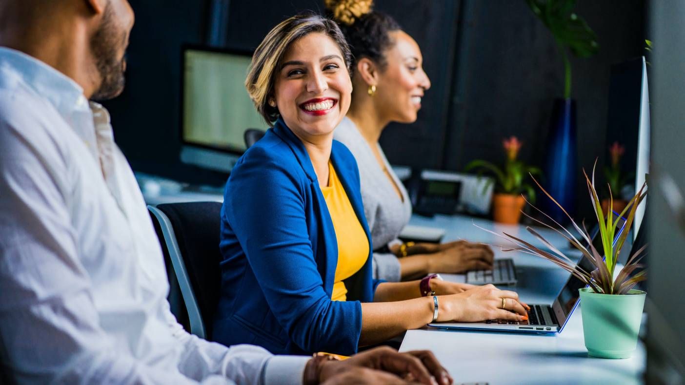 Smiling woman in blue blazer and yellow top working on laptop in a modern office.