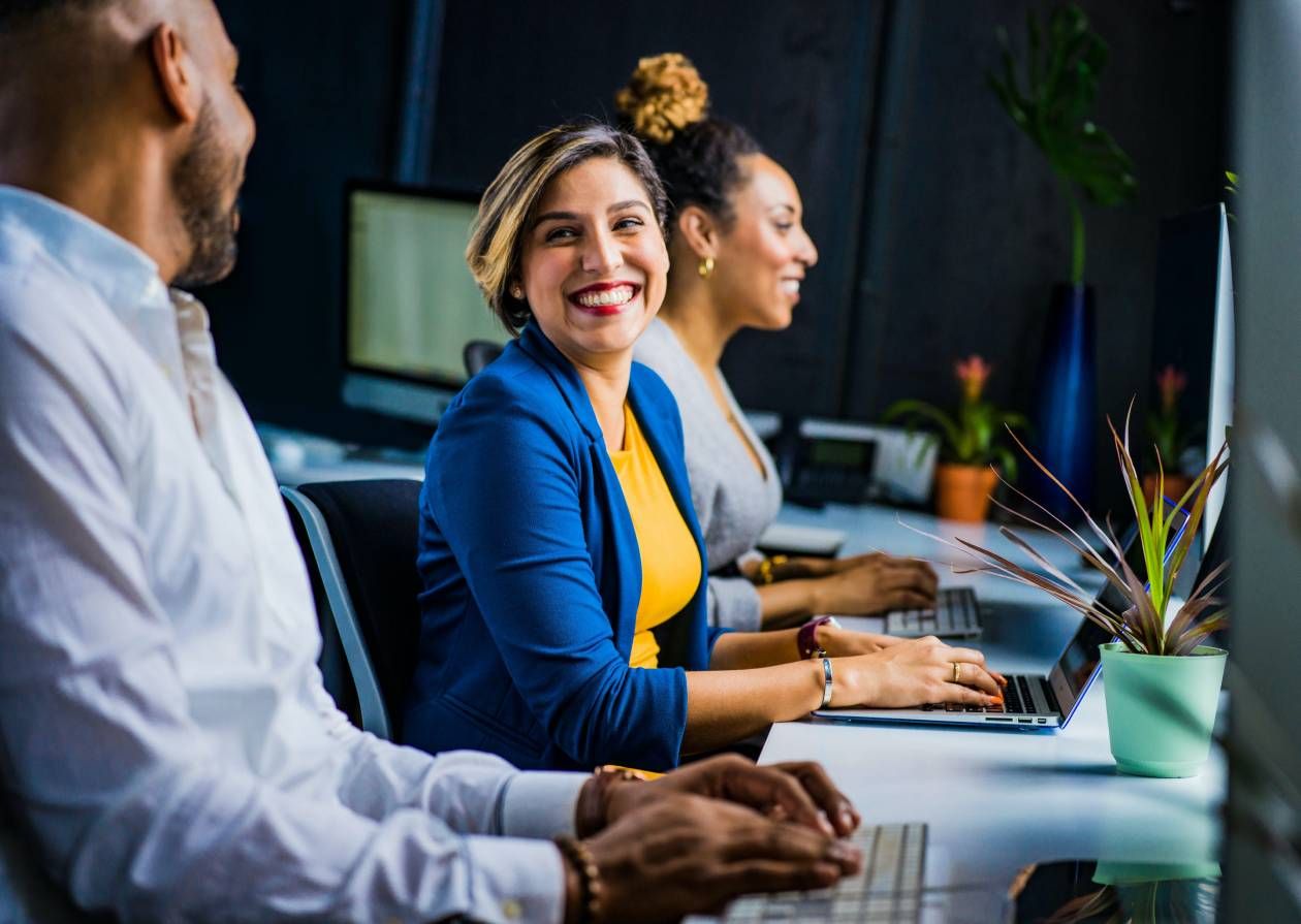 Three diverse professionals working at desks in an office, one woman smiling at the camera.