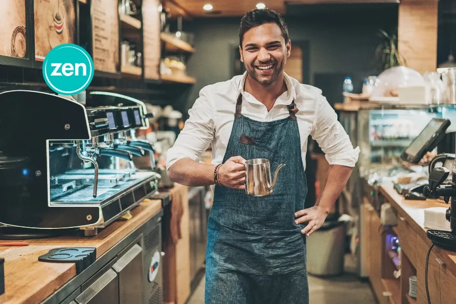 Smiling barista holding a coffee pitcher in a bustling coffee shop.