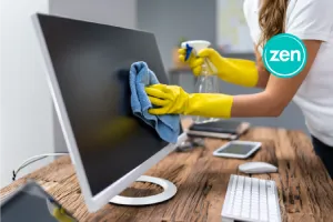 Person in yellow gloves cleans a computer monitor with a spray bottle and blue cloth.