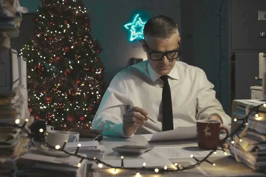 Man works late at a desk surrounded by Christmas decorations and paperwork.