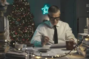 Man with glasses works at a cluttered desk decorated with Christmas lights and a tree.