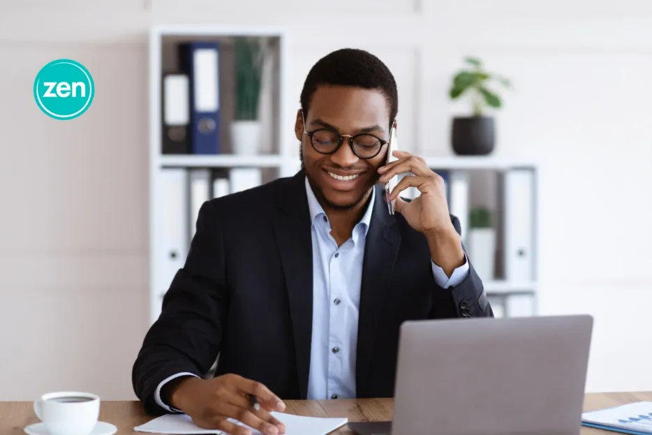 Smiling Black businessman in glasses talking on a phone at his office desk.