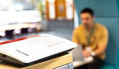 White and red router in foreground, blurred man using laptop in office background.