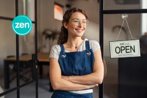 Smiling woman in an apron stands proudly by her shop's glass door with an 'OPEN' sign.