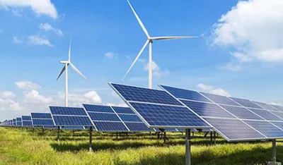 Solar panels in a green field with two large wind turbines under a blue sky.