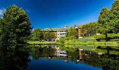 Large building with cupola reflected in a calm lake, surrounded by green trees under blue sky.
