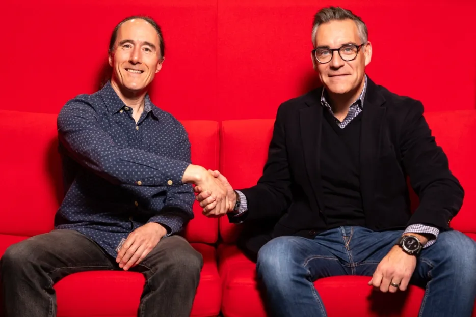 Two smiling men shaking hands on a vibrant red sofa, celebrating a partnership.