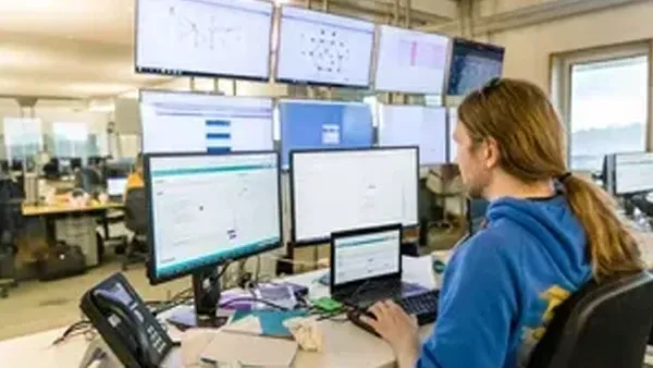 Person with long hair works at a desk surrounded by multiple computer monitors displaying data.