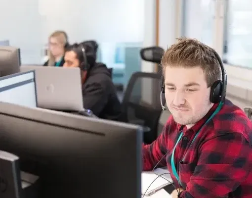 Man wearing a headset and red plaid shirt works at a computer in an office.