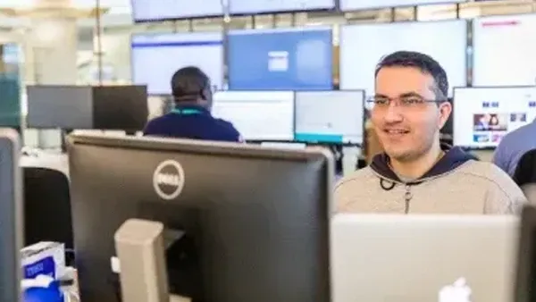 Man in glasses smiling at his computer in a modern office with multiple screens.