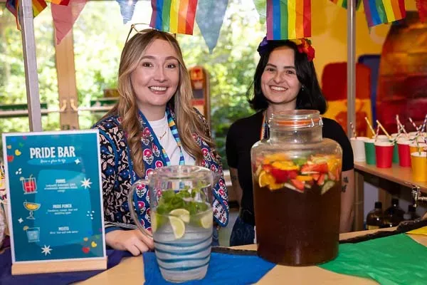 Two smiling women behind a Pride bar with fruit drinks and rainbow decorations.