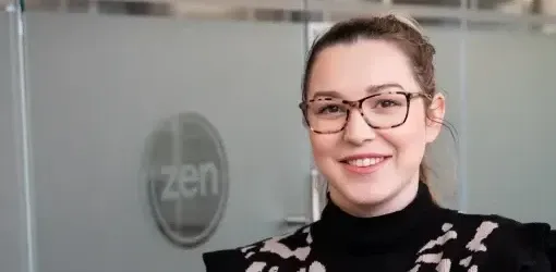 A young woman with glasses and a black patterned top smiles in an office.