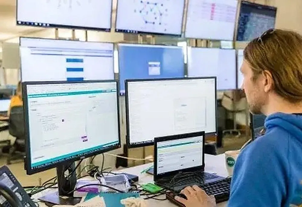 Person intensely works at a multi-monitor computer setup, analyzing data or code.