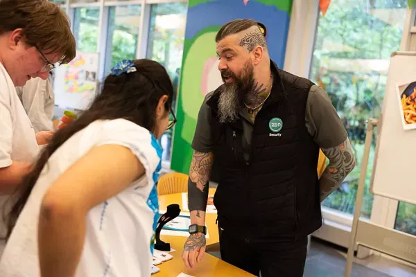 Bearded man with tattoos talking to a young girl in a classroom setting.